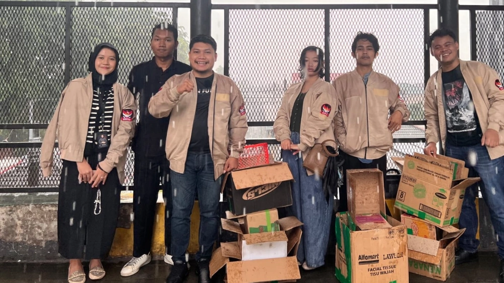 Pengurus dan kader DPP GEMPITA berjaket organisasi membawa kardus paket takjil di Halte Stasiun Cawang saat hujan